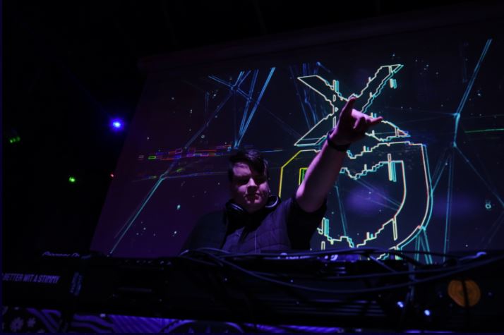 Image of electronic music producer xDiggityDee performing at a club with his xD logo behind him.